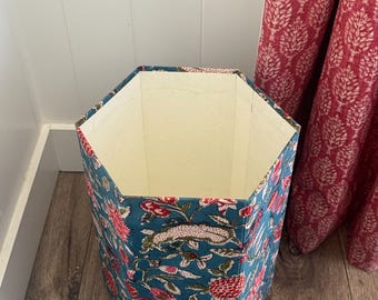 May include: A hexagonal wastebasket featuring a teal blue exterior with a repeating floral pattern in shades of pink, red, and green. The interior of the container is lined with a solid off-white fabric, and the structure has six distinct sides.