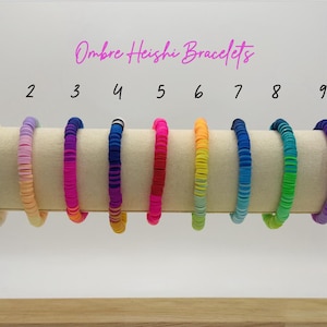 Op de afbeelding: Negen kleurrijke ombre heishi armbanden tentoongesteld op een beige cilindrische standaard. De armbanden zijn in een rij gerangschikt, elk met een unieke kleurovergang, variërend van lichtgroen tot paars. De tekst "Ombre Heishi Bracelets" staat erboven.