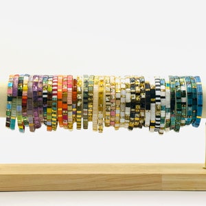 May include: A display of colorful beaded bracelets on a wooden stand. The bracelets are made of small, square beads in various colors and patterns.