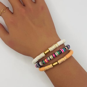 Puede incluir: Tres pulseras de cuentas de colores apiladas en una muñeca. Las pulseras presentan cuentas blancas, multicolores y color melocotón con detalles metálicos dorados. También se ve un anillo de corazón dorado.