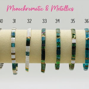 Op de afbeelding: Een rij van 11 armbanden met verschillende kleurcombinaties en patronen. De armbanden zijn gemaakt van kleine vierkante tegels in zwart, wit, goud en tinten blauw en groen. De armbanden zijn genummerd van 28 tot en met 38.