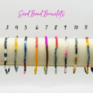 May include: A collection of thirteen seed bead bracelets in various colors, displayed on a white surface. Each bracelet features a unique combination of small, colorful beads. The text "Seed Bead Bracelets" is written above the bracelets.