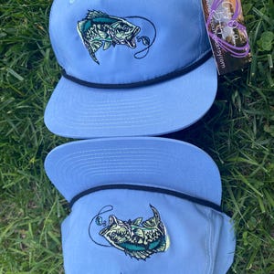 May include: Two light blue baseball caps with a black brim and a green and white embroidered fish design. The fish is depicted with a fishing hook in its mouth.