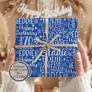 May include: A wrapped gift with blue wrapping paper featuring white text that says "Happy Birthday Eddie 70". The gift is tied with a gold ribbon. A circular graphic indicates custom name, age, and 15+ colors.