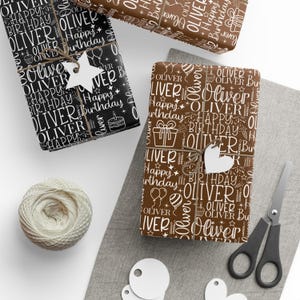 May include: Gift-wrapped presents with the name "Oliver" and "Happy Birthday" in white on black and brown wrapping paper. The gifts are tied with twine and adorned with star and heart-shaped tags. A ball of twine, scissors, and tags are also visible.