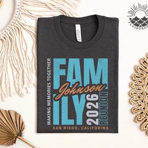 May include: Dark grey t-shirt with the words "FAMILY Johnson 2026 REUNION" in blue and orange. The text "MAKING MEMORIES TOGETHER" and "SAN DIEGO, CALIFORNIA" are also printed on the shirt. The shirt is folded on a white surface.