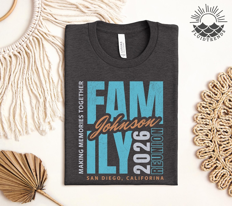 May include: Dark grey t-shirt with the words "FAMILY Johnson 2026 REUNION" in blue and orange. The text "MAKING MEMORIES TOGETHER" and "SAN DIEGO, CALIFORNIA" are also printed on the shirt. The shirt is folded on a white surface.