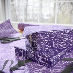 May include: Two wrapped gifts with purple wrapping paper featuring the name "Oliver" and "Happy Birthday" in white. The gifts are on a table with matching wrapping paper, scissors, and a roll of ribbon.