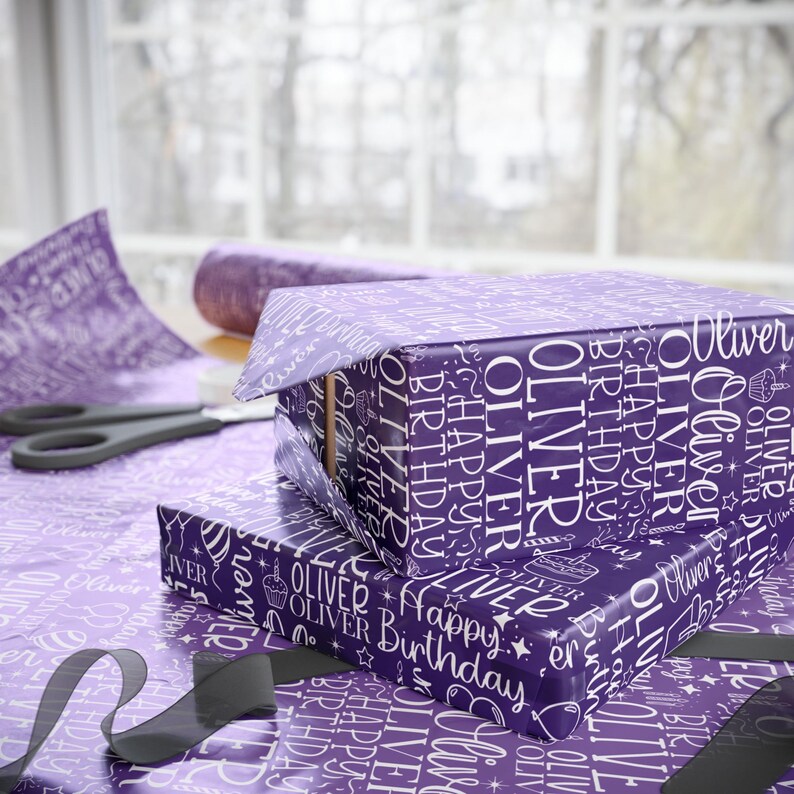 May include: Two wrapped gifts with purple wrapping paper featuring the name "Oliver" and "Happy Birthday" in white. The gifts are on a table with matching wrapping paper, scissors, and a roll of ribbon.