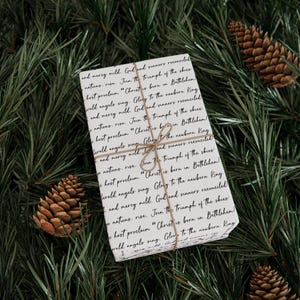May include: A wrapped gift with a white background and black script, tied with twine, rests on a bed of green pine needles. The gift is decorated with pine cones. The text on the gift reads "Christ is born in Bethlehem."