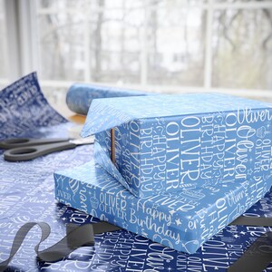 May include: Two gift boxes wrapped in light blue paper with white text that reads "Oliver" and "Happy Birthday." The boxes are stacked on a matching wrapping paper surface. Scissors and a roll of paper are in the background.
