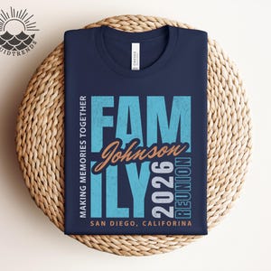 May include: A navy blue t-shirt featuring a teal and orange graphic that reads "FAMILY Johnson 2026 REUNION" and "MAKING MEMORIES TOGETHER" with "SAN DIEGO, CALIFORNIA" at the bottom. The shirt is folded on a woven surface.