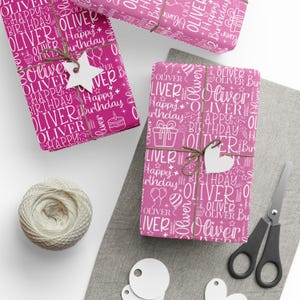 May include: Pink wrapping paper with a white pattern of the name "Oliver", birthday-themed icons, and the words "Happy Birthday". The wrapped gifts are tied with twine and adorned with white tags. A ball of twine and scissors are also present.