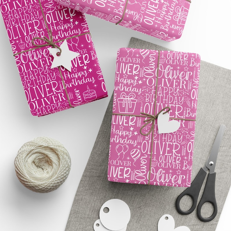 May include: Pink wrapping paper with a white pattern of the name "Oliver", birthday-themed icons, and the words "Happy Birthday". The wrapped gifts are tied with twine and adorned with white tags. A ball of twine and scissors are also present.