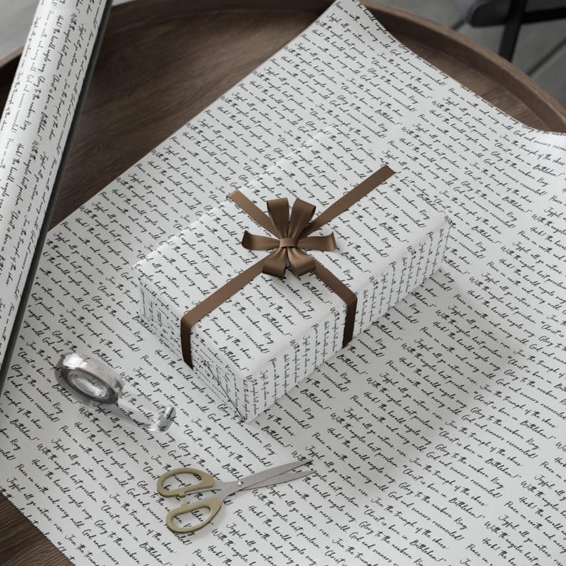 May include: White wrapping paper with black script, a wrapped gift with a brown ribbon, and a roll of paper. The gift is tied with a brown ribbon. Scissors and tape are also visible. The paper has text that reads "Glory to the newborn king!"