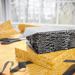 May include: Two wrapped gift boxes, one black and one yellow, with the name "Oliver" and "Happy Birthday" printed on them. A pair of scissors and a roll of wrapping paper are visible in the background. A black ribbon is also present.