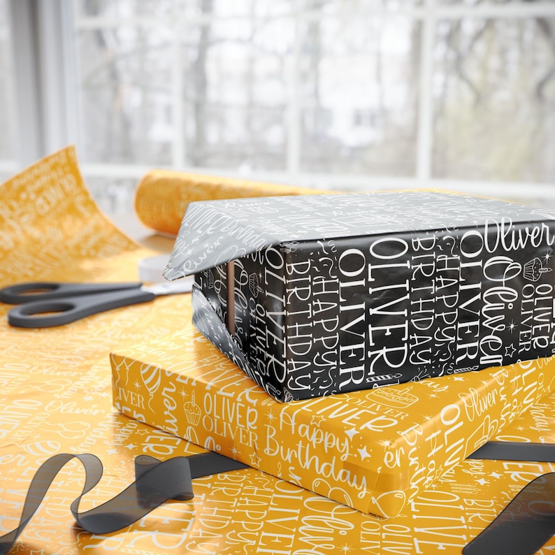 May include: Two wrapped gift boxes, one black and one yellow, with the name "Oliver" and "Happy Birthday" printed on them. A pair of scissors and a roll of wrapping paper are visible in the background. A black ribbon is also present.