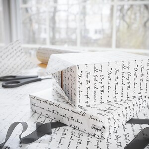 May include: White gift boxes wrapped in paper with black script. A roll of matching paper and black ribbon are nearby. Scissors are also visible. The wrapping paper has a handwritten script design.