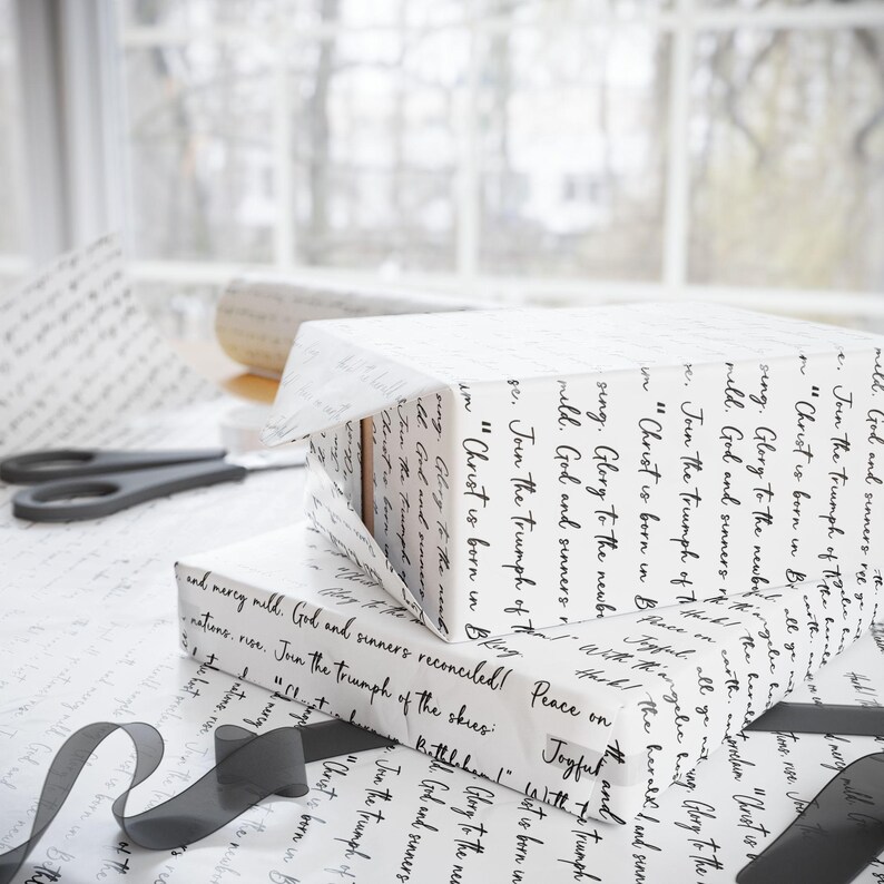 May include: White gift boxes wrapped in paper with black script. A roll of matching paper and black ribbon are nearby. Scissors are also visible. The wrapping paper has a handwritten script design.