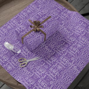 May include: Purple wrapping paper with a repeating pattern of the name "OLIVER" and "Happy Birthday" in white. A wrapped gift with a brown bow, scissors, and tape are on top of the paper. A festive gift wrapping theme.