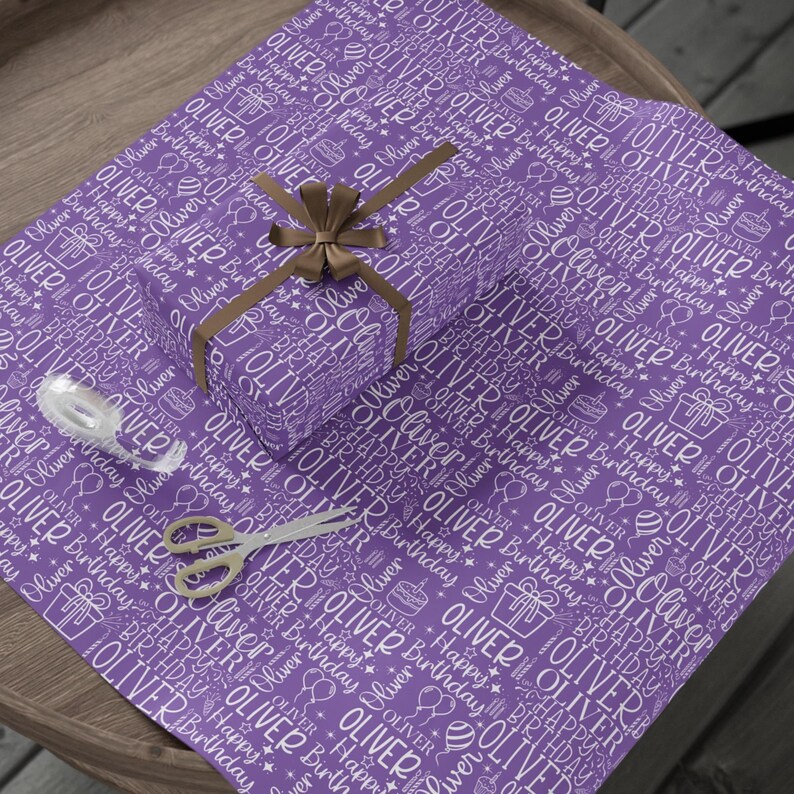 May include: Purple wrapping paper with a repeating pattern of the name "OLIVER" and "Happy Birthday" in white. A wrapped gift with a brown bow, scissors, and tape are on top of the paper. A festive gift wrapping theme.
