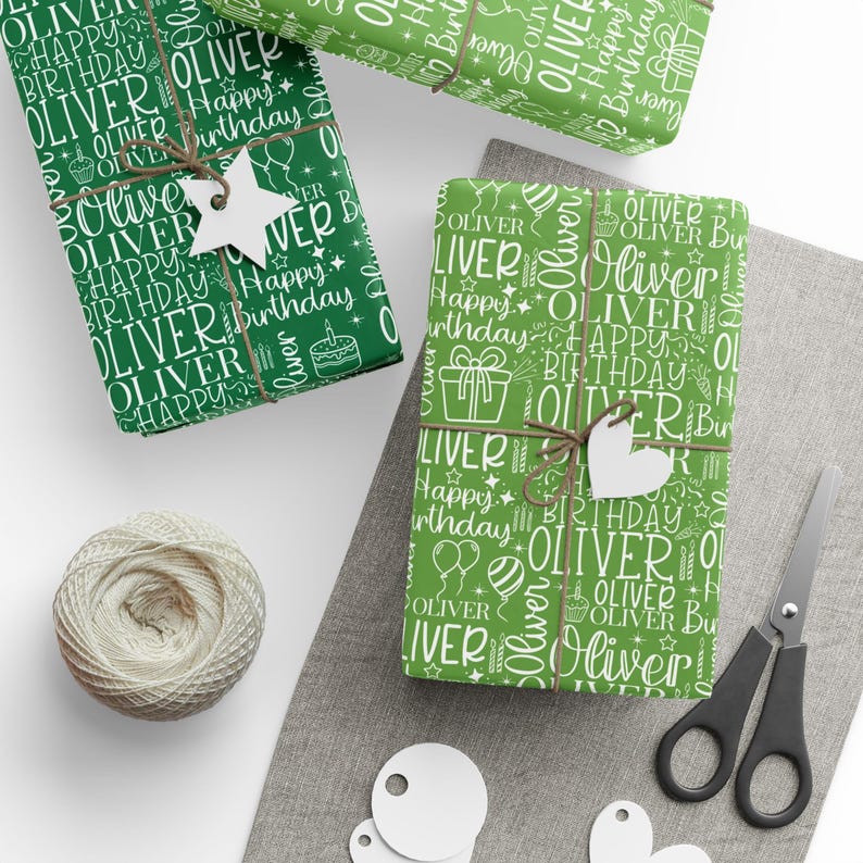 May include: Green wrapping paper with white text that says "Oliver Happy Birthday" is wrapped around gifts. The gifts are tied with twine and adorned with white star and heart-shaped tags. A spool of twine and scissors are also present.