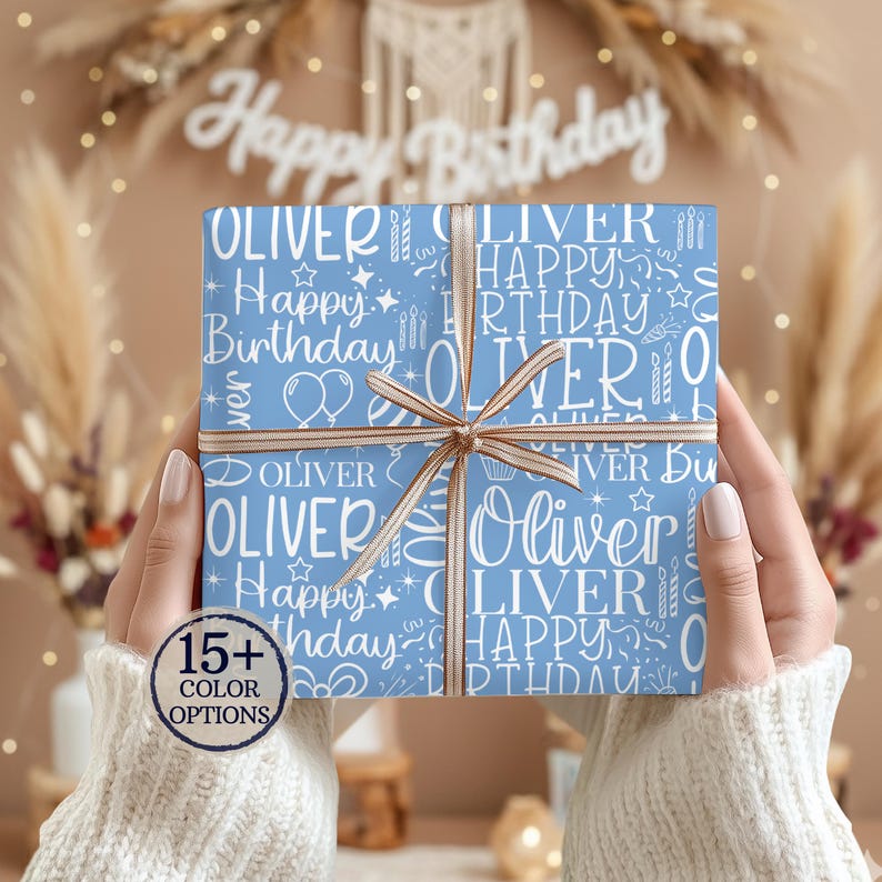 May include: A wrapped gift with a light blue background featuring the name "Oliver" and the words "Happy Birthday" in white. The gift is tied with a brown ribbon. The words "15+ COLOR OPTIONS" are in a circle in the lower left corner.