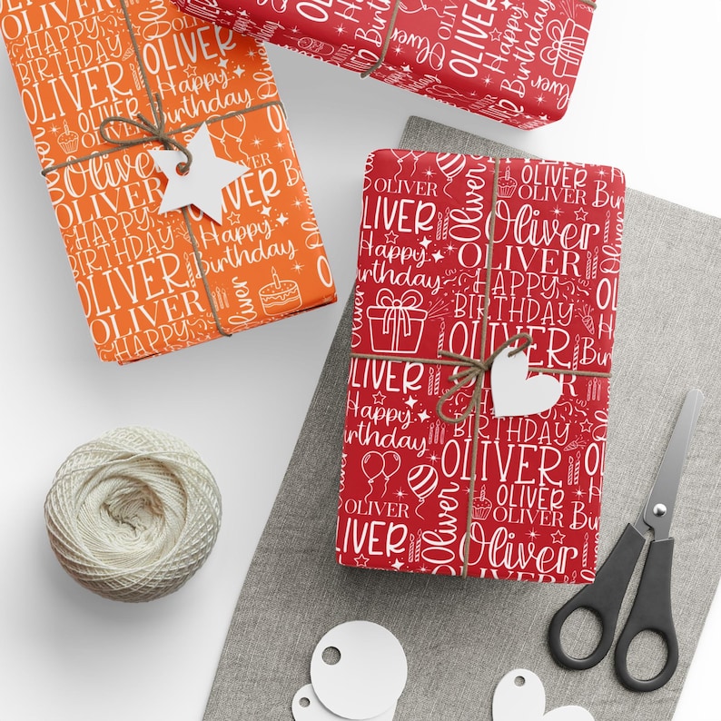 May include: Three wrapped gifts with birthday-themed wrapping paper. The paper is red and orange with the name "Oliver" and "Happy Birthday" in white. Each gift is tied with twine and a tag. A ball of twine, scissors, and tags are also present.