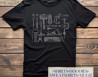 Op de afbeelding: Een zwart t-shirt met korte mouwen en een witte lijntekening van diverse houtbewerkingsgereedschappen, waaronder een hamer, zaag, schaaf en beitels. Een rechthoekig label in de rechterbenedenhoek toont de tekst SHIRTS HOODIES SWEATSHIRTS MUGS.