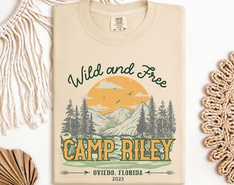 May include: A beige t-shirt with a green and yellow graphic design. The graphic features a mountain range with a sunset, trees, and the text "Wild and Free Camp Riley Oviedo, Florida 2023".