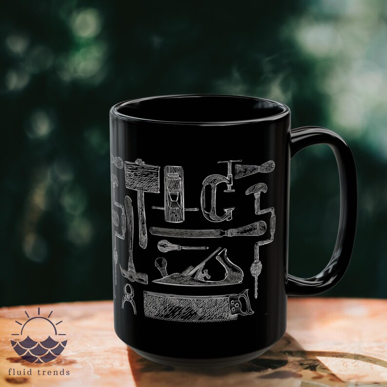 May include: Black ceramic mug with a white illustration of carpentry tools, including a hammer, saw, and plane. The mug has a large handle and a glossy finish. The mug is displayed on a wooden surface. The mug is a great gift for a carpenter.