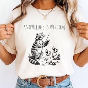 May include: Cream-colored t-shirt featuring a black and white illustration of a raccoon teaching two smaller raccoons. The text "KNOWLEDGE IS WISDOM" is above the illustration. The shirt has short sleeves and a crew neck.