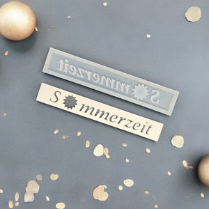 Könnte beinhalten: Zwei rechteckige Holzschilder mit einem Sonnenmotiv und dem Text "Sommerzeit" in weißen Buchstaben. Die Schilder befinden sich auf einem grauen Hintergrund mit kleinen weißen und braunen Punkten.