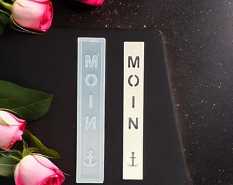 May include: Two white rectangular bookmarks with the word "MOIN" and an anchor symbol. The bookmarks are on a black surface with pink roses.