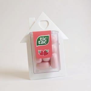 Könnte beinhalten: Ein weißer, hausförmiger Halter mit einem transparenten Kunststoffbehälter mit rosa Tic Tac-Bonbons. Ein rotes Etikett mit dem Tic Tac-Logo und Erdbeer-Grafik ist angebracht.