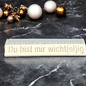 Könnte beinhalten: Eine weiße rechteckige Silikonform mit dem Text "Du bist mir wichtig(el)ig" geprägt auf der Oberfläche. Die Form liegt auf einer schwarzen Marmorfläche mit goldenen und weißen Ornamenten.