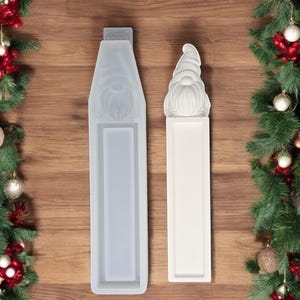 Könnte beinhalten: Zwei weiße Silikonformen zum Herstellen von gnomförmigen Dekorationen. Die Formen sind rechteckig und haben ein erhabenes Design eines Gnomkopfes und Bartes.