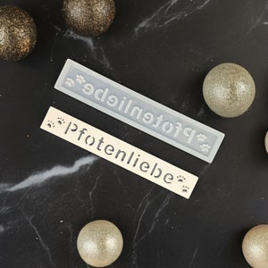 Könnte beinhalten: Zwei weiße rechteckige Schilder mit dem Text "Pfoetenliebe" in schwarzer Schrift und Pfotenabdrücken. Die Schilder liegen auf einer schwarzen Marmorfläche mit goldenen und silbernen Ornamenten.