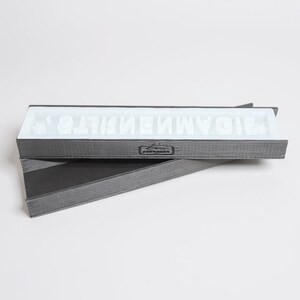 Könnte beinhalten: Eine graue rechteckige Box mit weißem Innenraum enthält weiße Buchstaben, die ein Wort bilden. Die Box ist geöffnet und zeigt die Buchstaben im Inneren. Die Box besteht aus dunklem grauem Material und steht vor weißem Hintergrund.