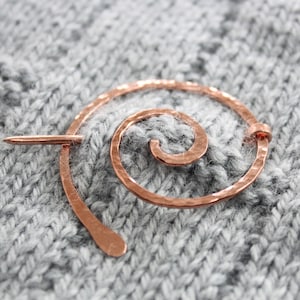 Peut inclure: Un porte-aiguilles à tricoter en spirale de couleur cuivre avec une finition texturée. Le porte-aiguilles est sur un tissu tricoté gris.
