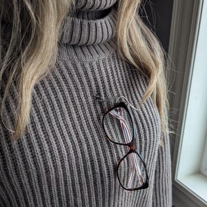 May include: A pair of spectacles with a brown frame and pink accents are hanging from a grey sweater.