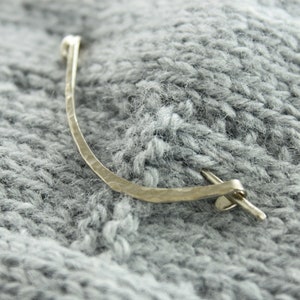 May include: A silver metal hair clip with a hammered finish and a simple design. The clip is lying on a gray knitted fabric.