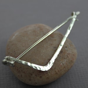 May include: A silver hammered metal brooch in the shape of a triangle with a straight bar across the top. The brooch is resting on a brown rock.