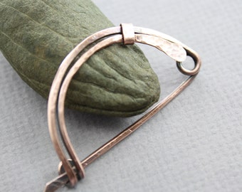 May include: A copper brooch with a hammered finish and a curved shape. The brooch is made of two pieces of copper wire that are twisted together.