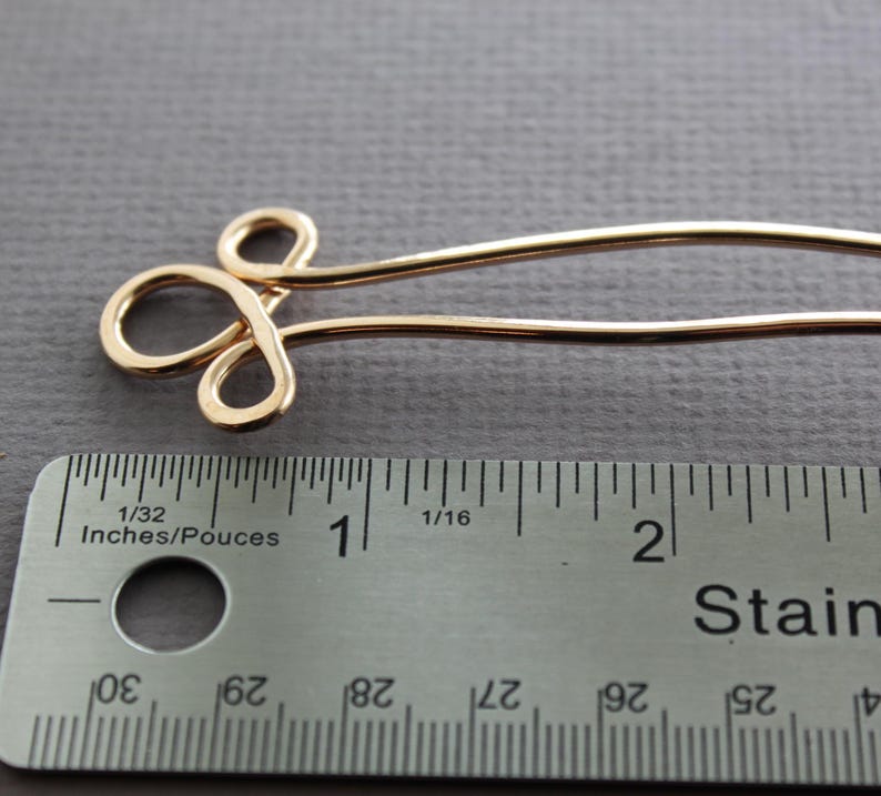 May include: Gold-toned metal hair pin with a decorative loop design. The pin is approximately 1 inch long and is measured against a ruler.