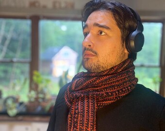 Könnte beinhalten: Ein Mann trägt ein schwarzes T-Shirt und einen schwarz-orangen gestreiften Schal. Er trägt schwarze Kopfhörer und schaut nach rechts.