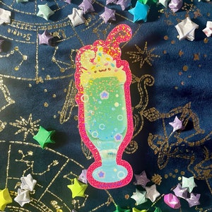 May include: A pink and green glitter milkshake with a white and yellow cartoon character on top. The milkshake is in a clear glass with a pink rim and is surrounded by origami stars.