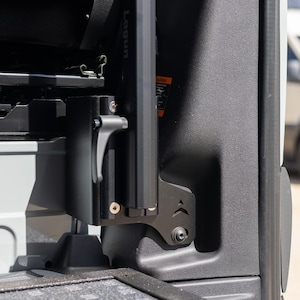 May include: Close-up of a black metal component with a silver handle, mounted on a gray interior panel. The component appears to be part of a vehicle's interior, possibly a seat mechanism. A white van is visible in the background.