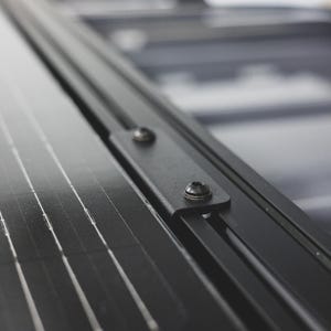 May include: A black solar panel mounted to a black metal roof rack system. The panel features a grid of thin silver conductive lines and is secured to the frame with a black metal bracket held in place by two visible silver-toned metal screws.