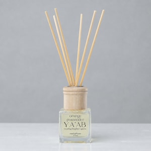 May include: A clear glass reed diffuser with a light wood collar and six light-colored wooden reeds. The label reads orange pomander YA'AB creating brighter spaces reed diffuser 3.70oz. The bottle is square-shaped with a thick base.